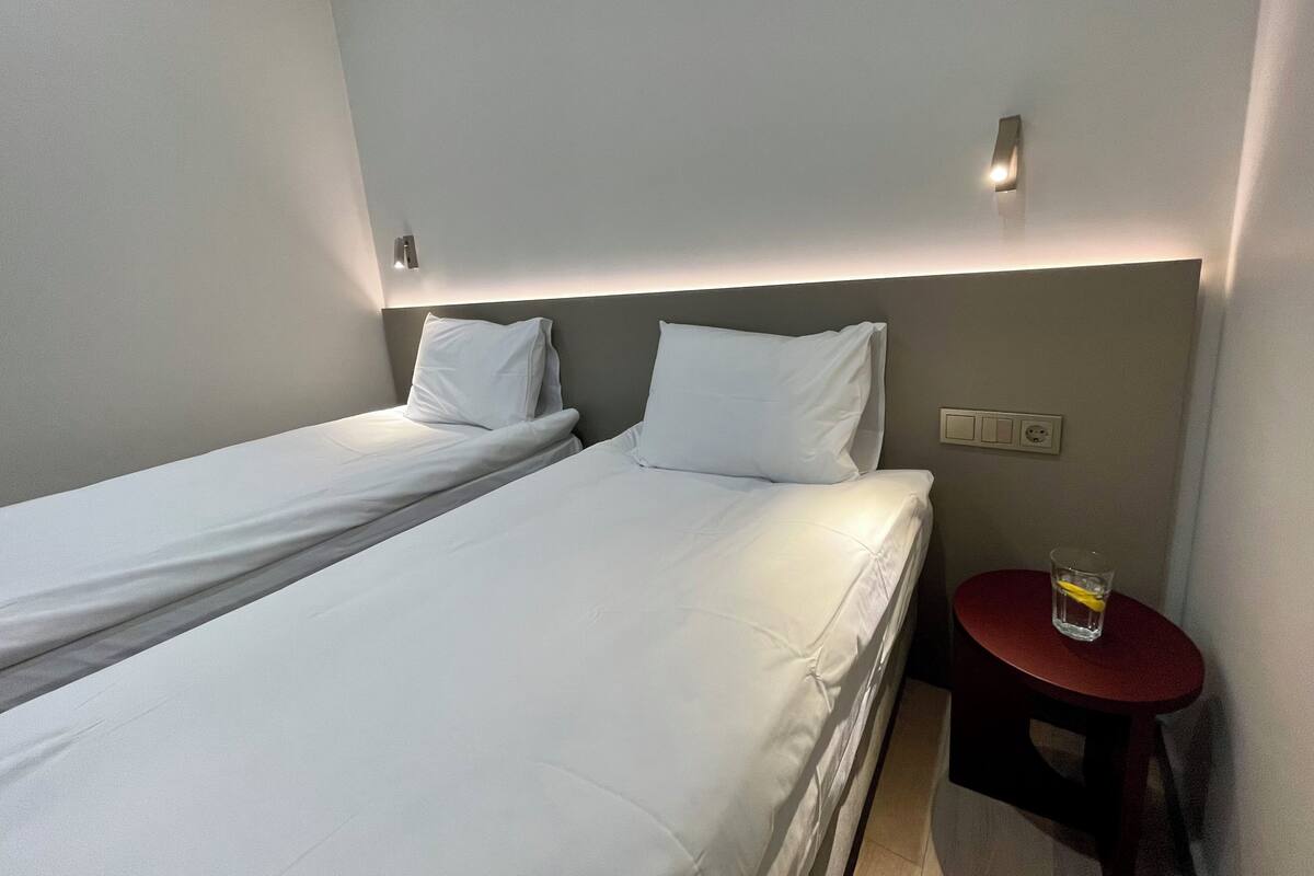 A hotel room with two single beds, a small round table with a glass of water, and a wall socket.