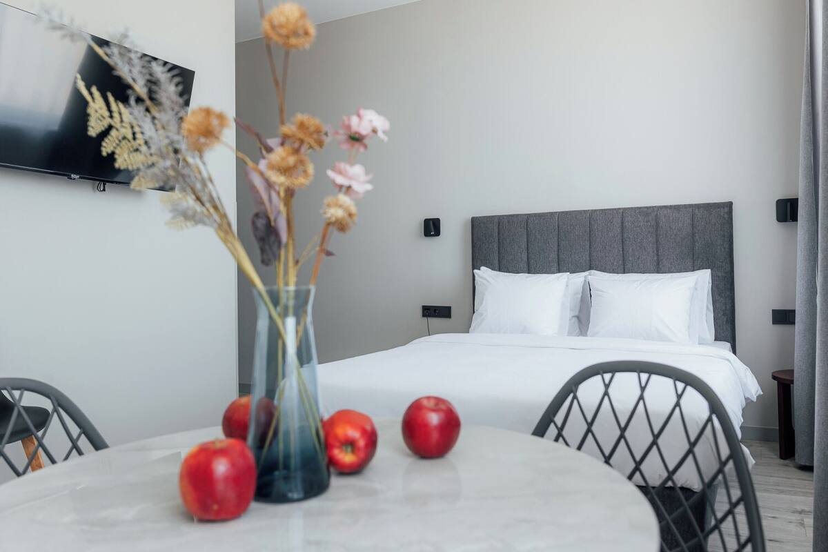 A modern hotel room with a bed, a TV mounted on the wall, a vase with dried flowers, and a table with apples.