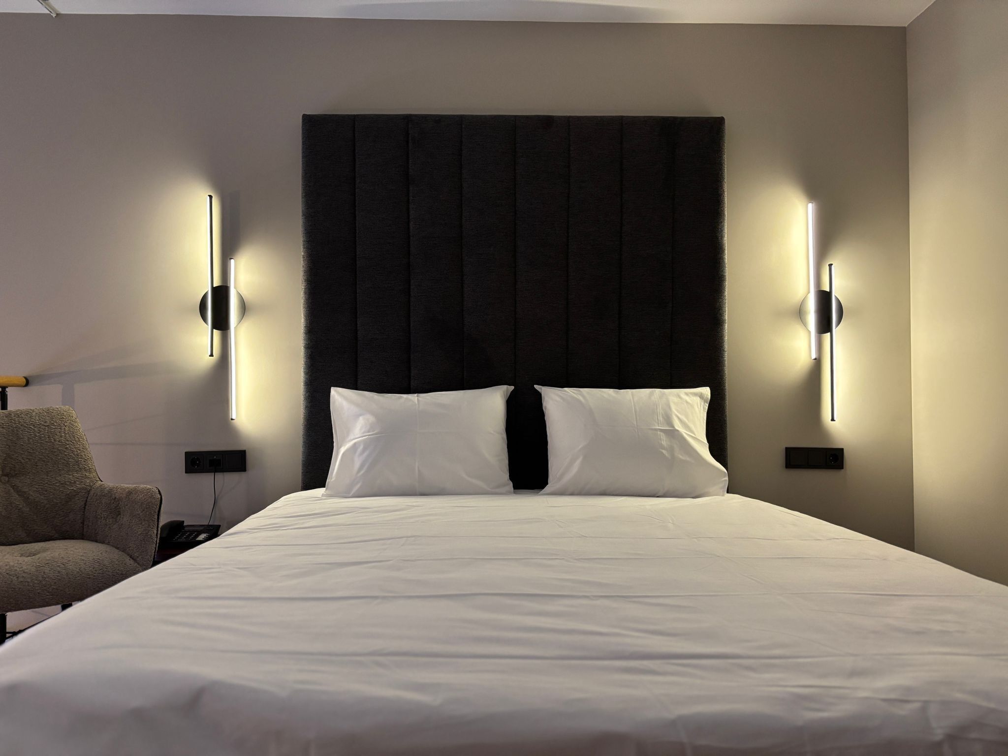 A hotel room with a large bed, two bedside tables, a chair, and modern wall sconces.