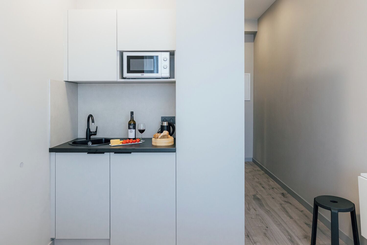 Una kitchenette compacta con microondas, lavamanos y encimera con alimentos y una copa de vino.