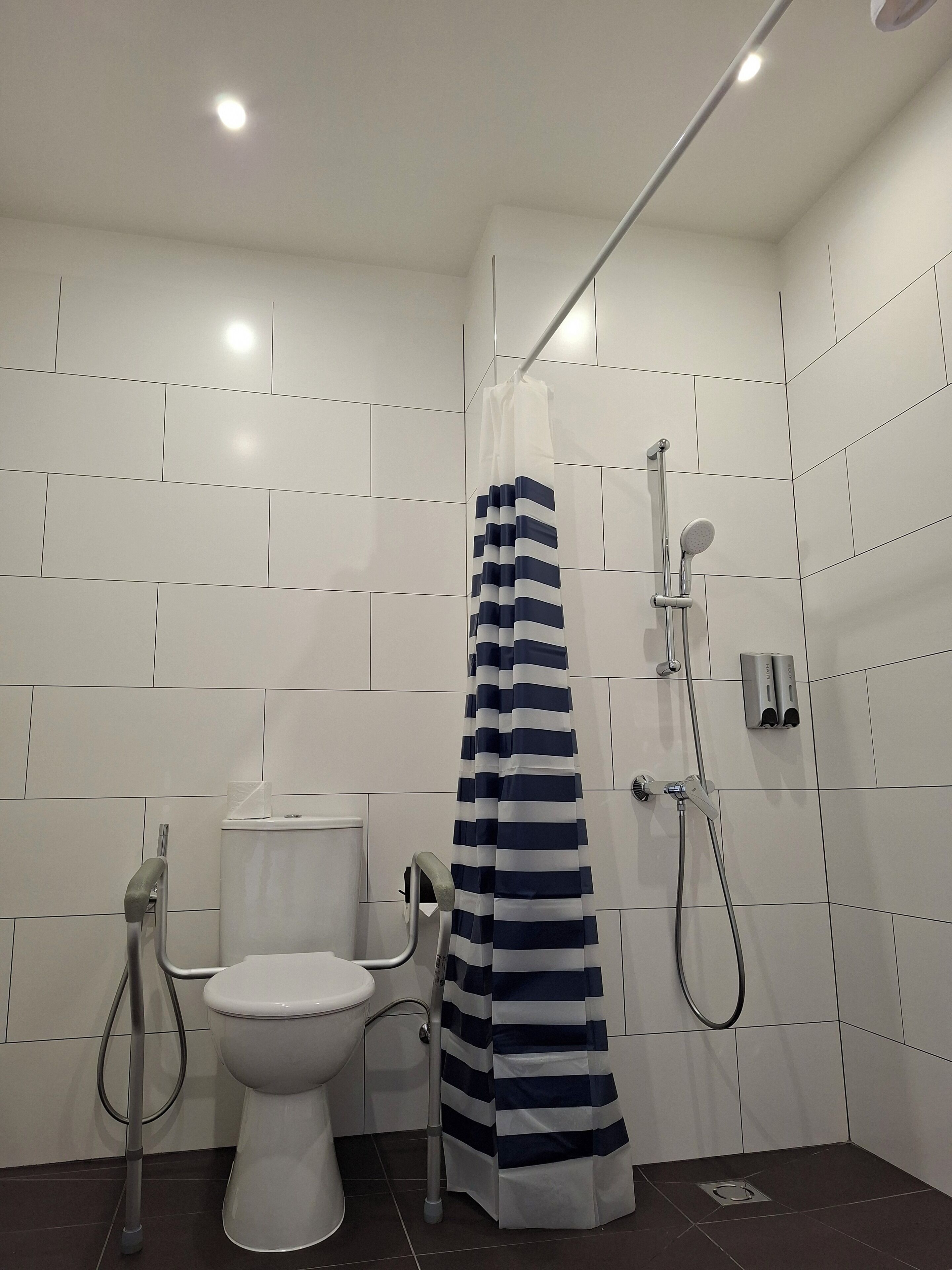 Ett modernt badrum med vita kakelplattor, en vit toalett, en dusch med ett randigt duschdraperi och ett duschmunstycke som kan hållas i handen.