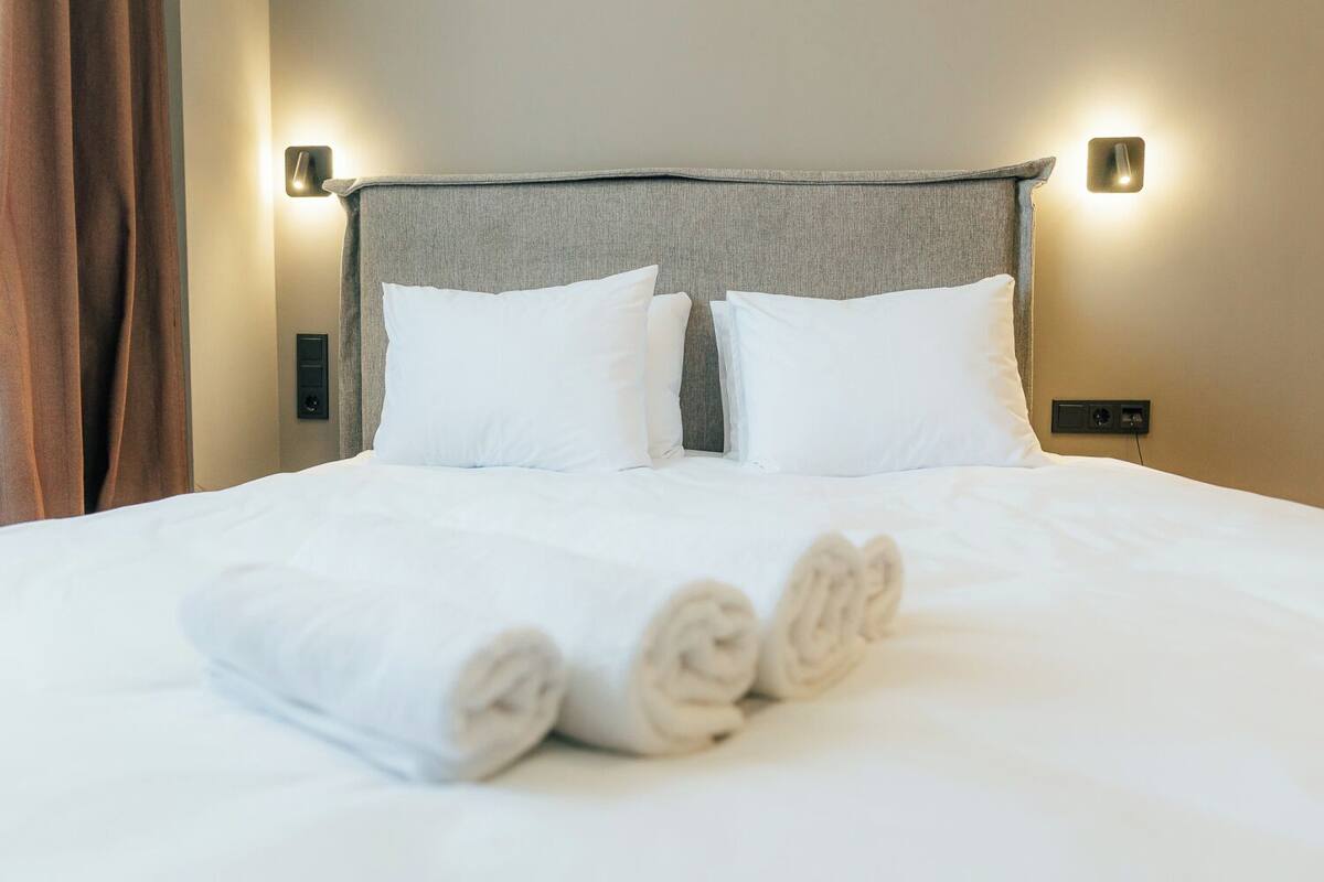 A neatly made bed with white linens and rolled towels, a grey headboard, and two wall-mounted light fixtures.