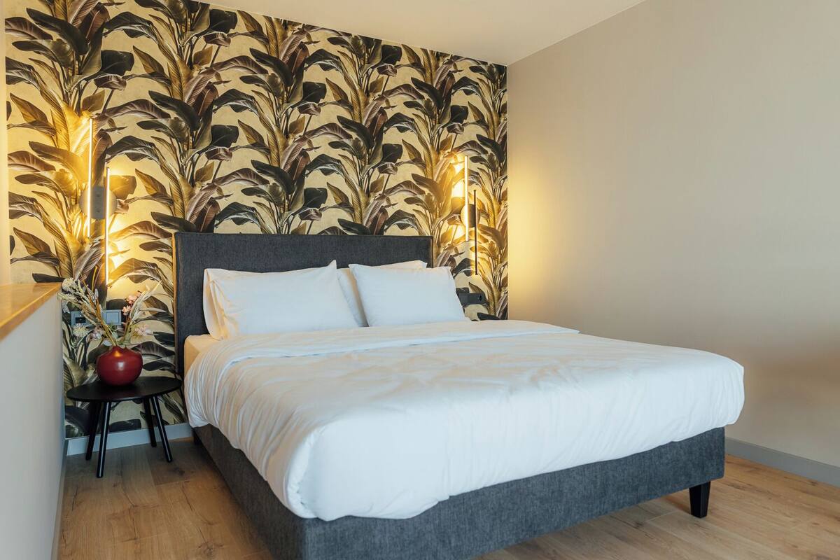 A neatly made bed with white bedding, a dark grey headboard, and a wall with a leafy pattern.