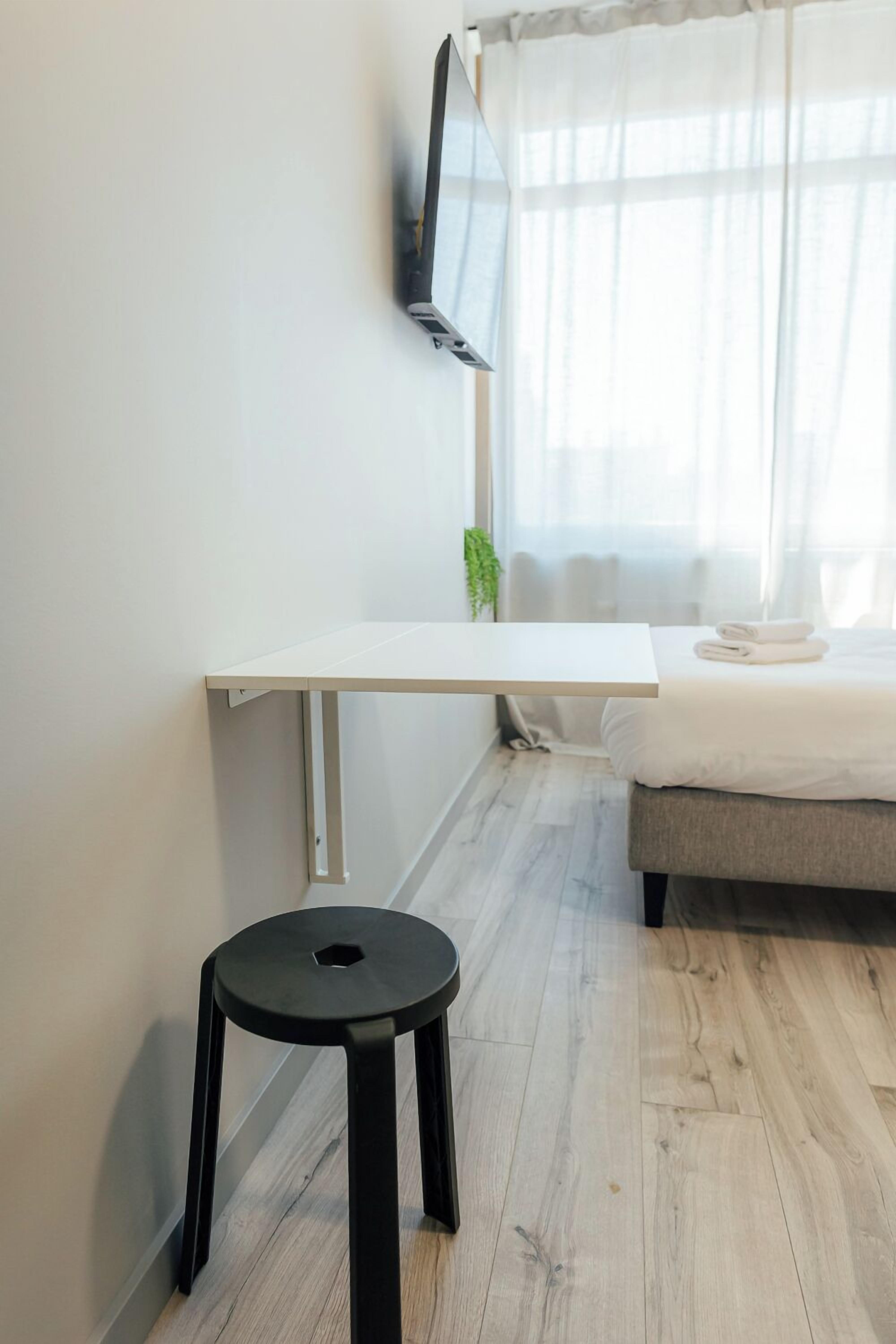 Een kamer met een wit bureau, een zwarte stoel, een bed met witte beddengoed, een aan de muur bevestigde televisie en een raam met dunne gordijnen.