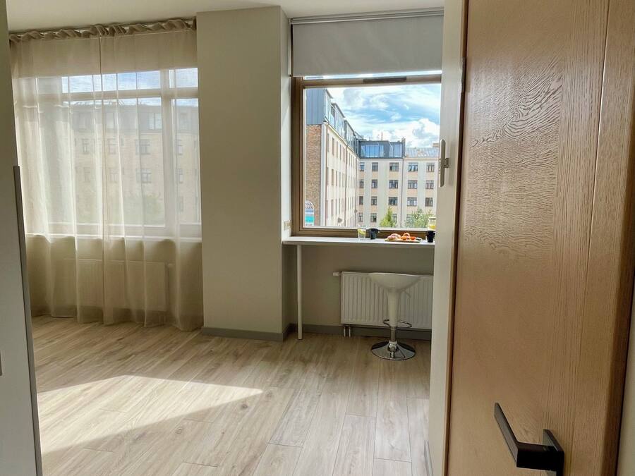 Una habitación con suelo de madera, ventana con vista a edificios, un radiador y una puerta con manija.