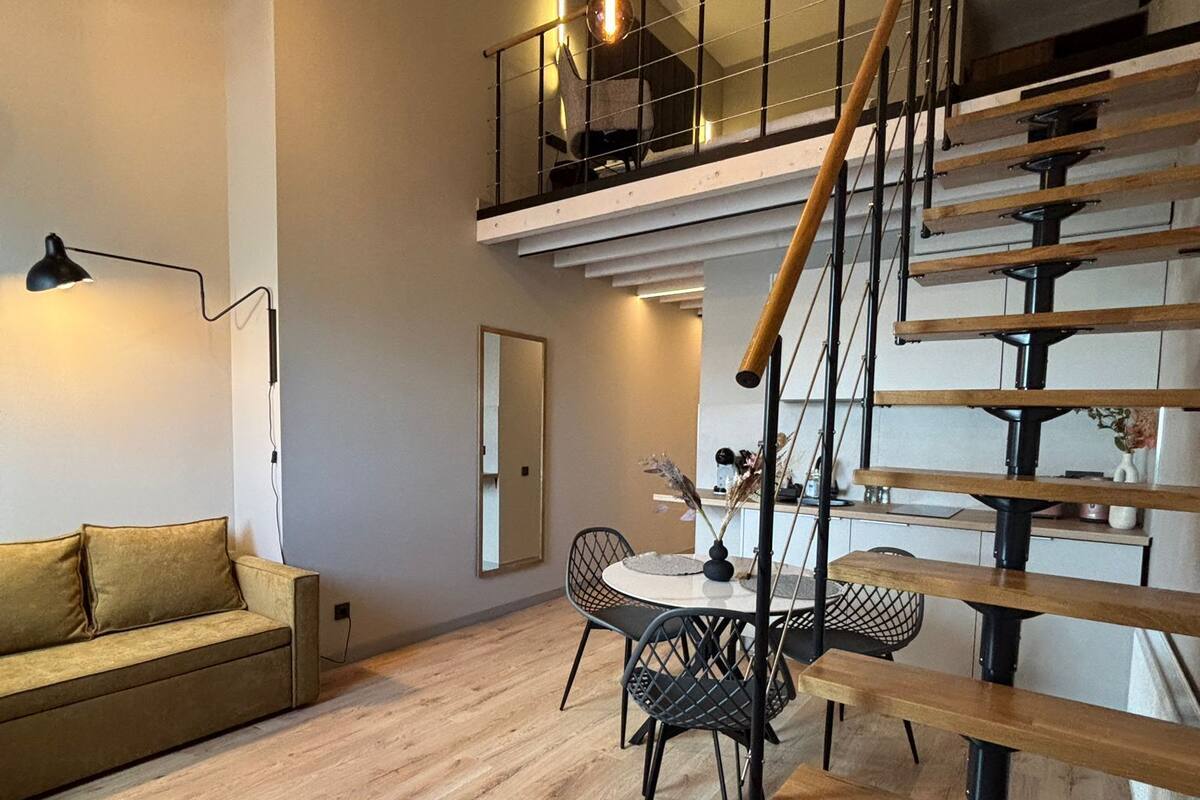 A modern loft-style apartment with a wooden staircase, a dining area, and a living room with a sofa and a lamp.