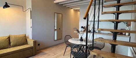 A modern loft-style apartment with a wooden staircase, a dining area, and a living room with a sofa and a lamp.