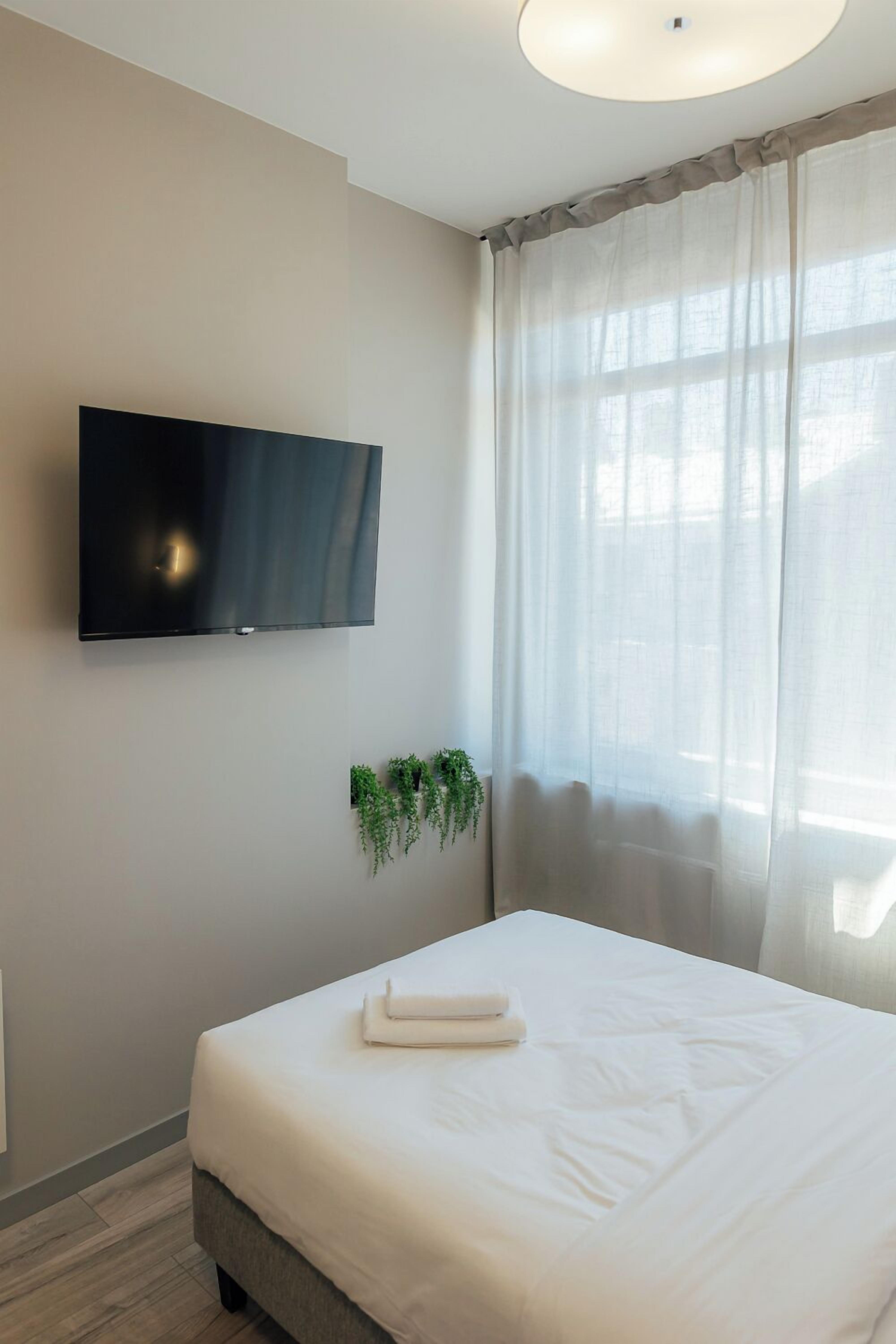 Een hotelkamer met een bed, een wandtelevisie, een raam met dunne gordijnen en een kleine plant op de vensterbank.