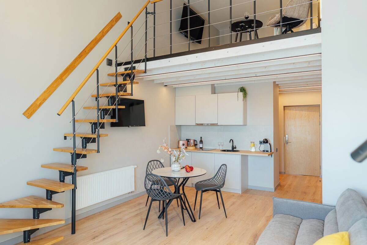 A modern apartment with a wooden staircase, a dining area with a round table and chairs, a kitchen with white cabinets, and a living area with a sofa.