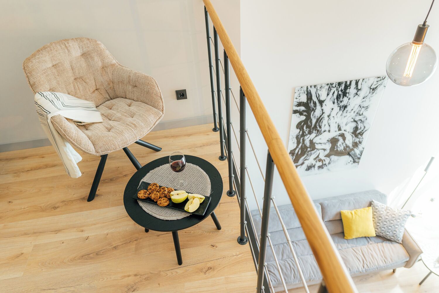 Uma sala de estar moderna com piso de madeira, uma mesa de centro redonda preta com petiscos e um copo de vinho tinto, uma poltrona bege, um sofá cinza com uma almofada amarela e um corrimão de escada em metal preto.