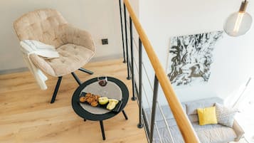 A modern living room with a wooden floor, a round black coffee table with snacks and a glass of red wine, a beige armchair, a grey sofa with a yellow pillow, and a black metal staircase railing.