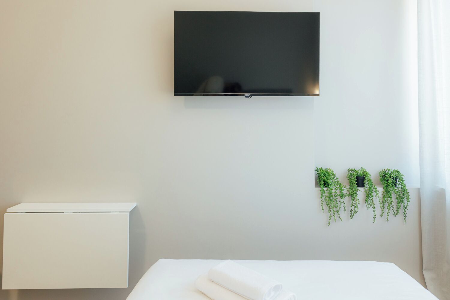 A hotel room with a flat-screen TV mounted on the wall, a white bedside table, a white bed with a neatly folded white towel, and three hanging green plants.