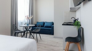 A modern hotel room with a bed, a dining area with a table and chairs, a sofa, a TV mounted on the wall, and a small kitchen area with a kettle and plant. - Aparthotel Aurum (Rīga)