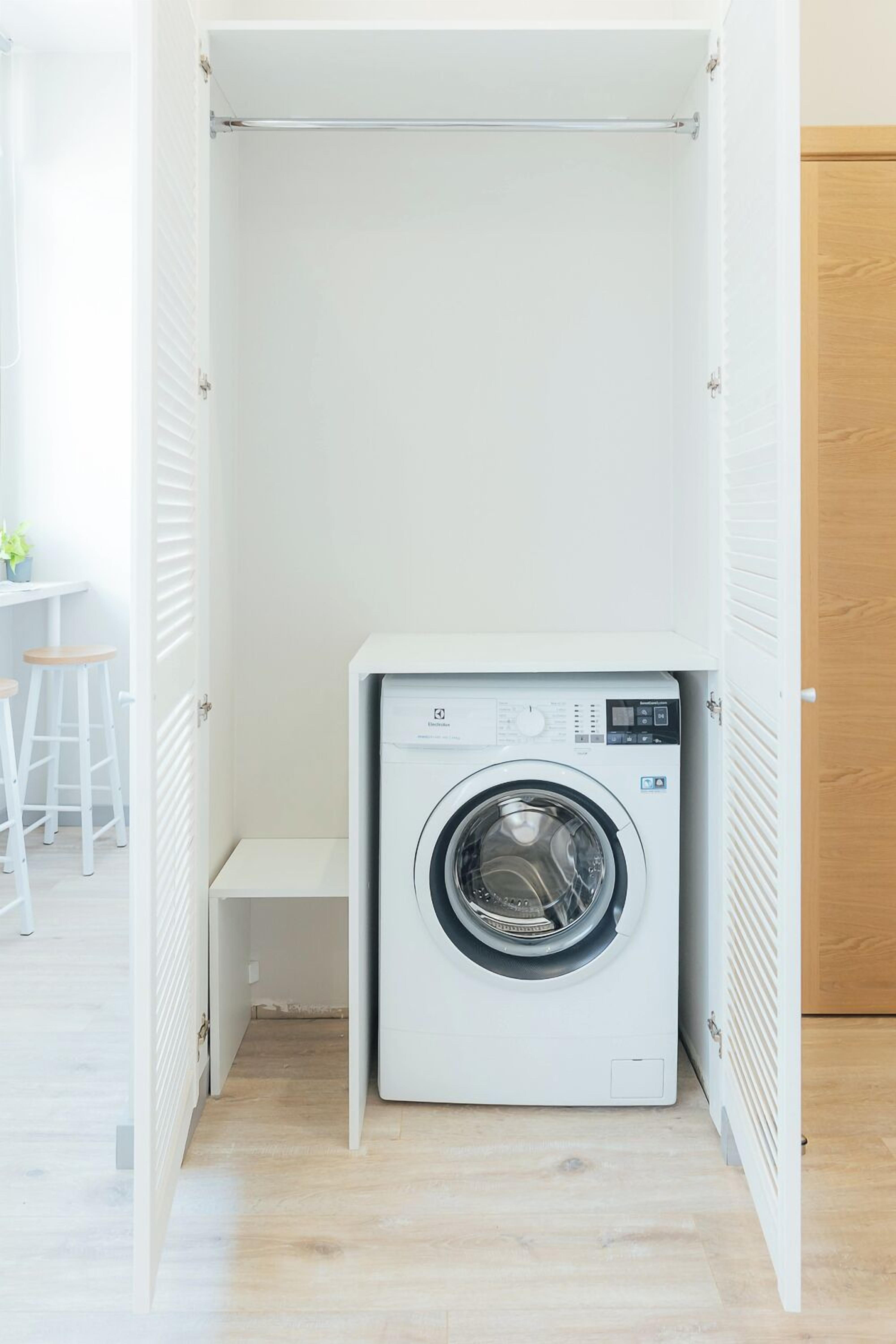 A compact, white laundry room with a front-loading washing machine, a bench, and a small table with stools.