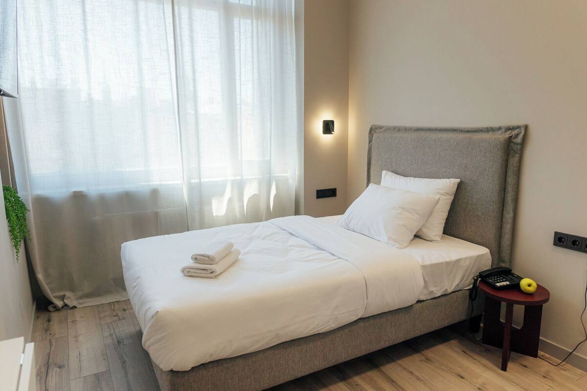 A neatly made bed with white linens and a folded towel, a bedside table with a phone and an apple, and a wall-mounted light.