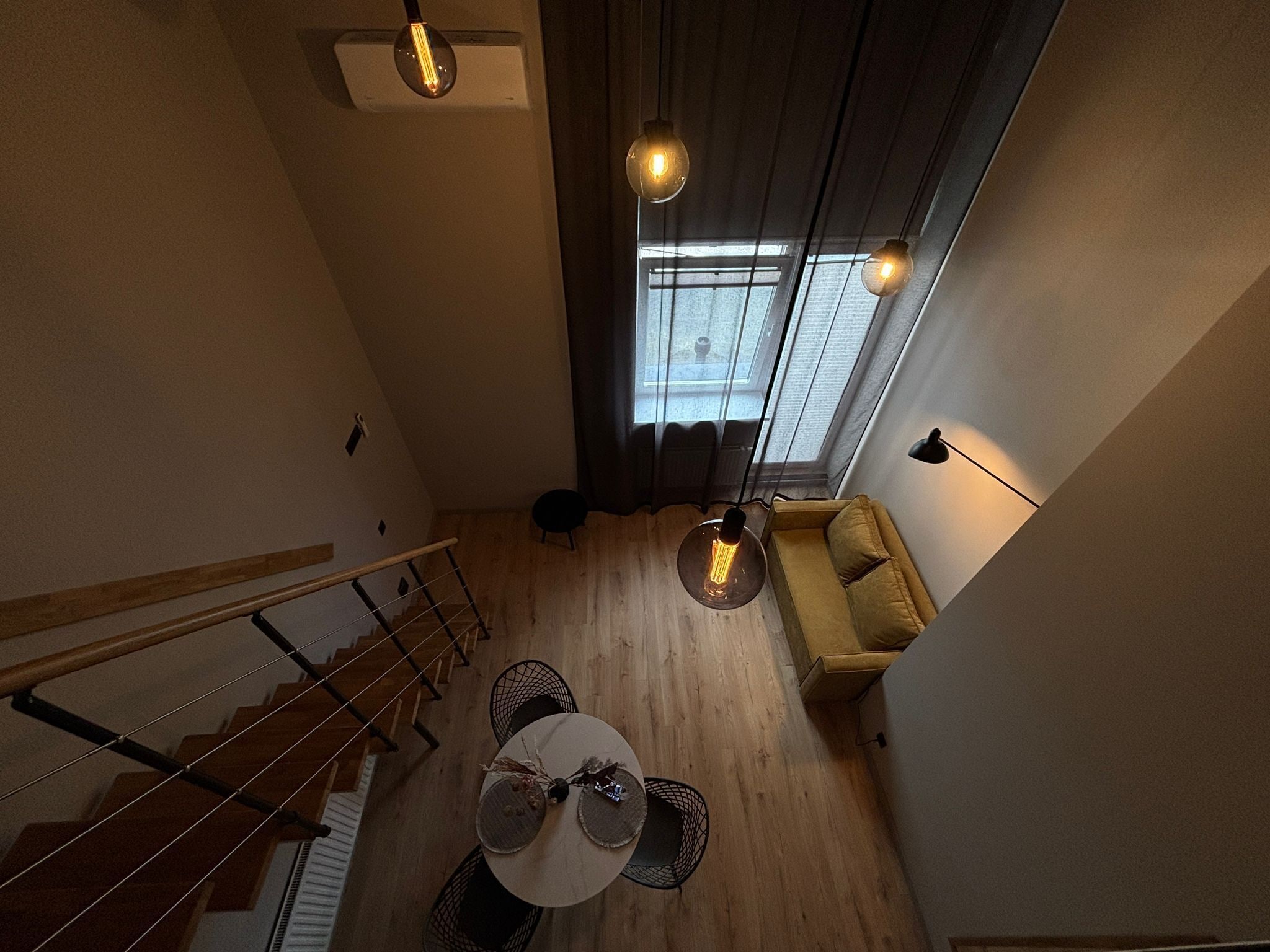 A high-ceilinged room with a staircase, a round table with a vase, and hanging pendant lights.