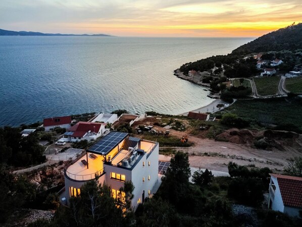 Una propiedad costera con una casa moderna, de techo curvo y paneles solares, con vistas a un mar tranquilo y un paisaje montañoso.