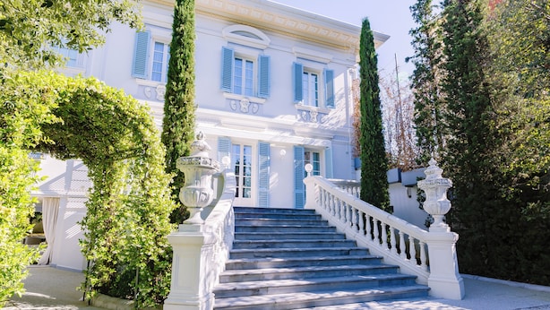 Una imponente escalera conduce a un edificio blanco con contraventanas azules, rodeado de abundante vegetación.