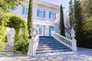 A grand staircase leading to a white building with blue shutters, surrounded by lush greenery.