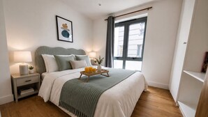 A bedroom with a bed, bedside tables, a window with curtains, and a view of buildings outside.