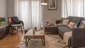 A modern living room with a sofa, a coffee table, and a chair. There is a lamp, a framed picture on the wall, and a window with sheer curtains. - Calma Center Granada (Granada)