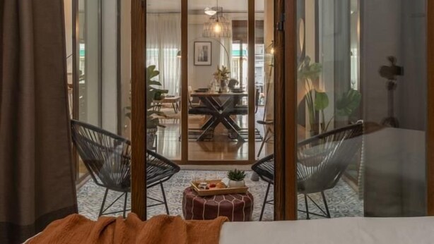 A modern hotel room with a large sliding glass door leading to a dining area. There is a bed with a blanket, a small table with a tray of snacks, and two black chairs. - Calma Center Granada (Granada)