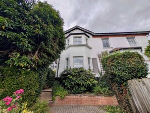 A two-story building with a white exterior, a brick pathway leading to the entrance, and dense greenery surrounding the property. - Charming Two-Bedroom House in London (Greater London)
