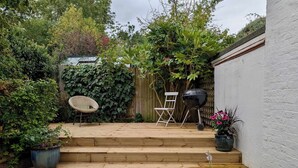 Eine Holzterrasse mit Stufen, die zu einer Sitzgelegenheit führen, umgeben von üppigem Grün und einer weißen Mauer.