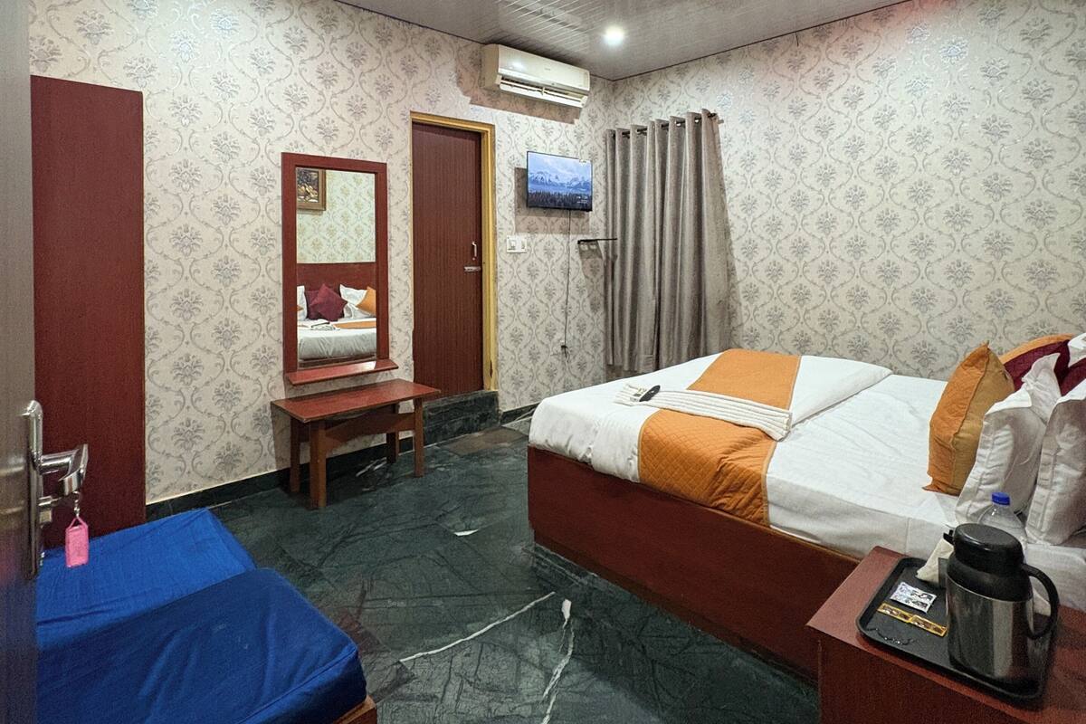 A hotel room with a bed, a desk, a chair, a mirror, a television, and an air conditioning unit.