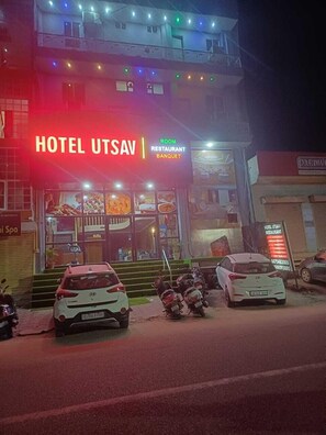 A building with a sign reading "HOTEL UTSAV RESTAURANT BANQUET" and multiple parked vehicles in front. - Hotel Utsav (Jaipur)
