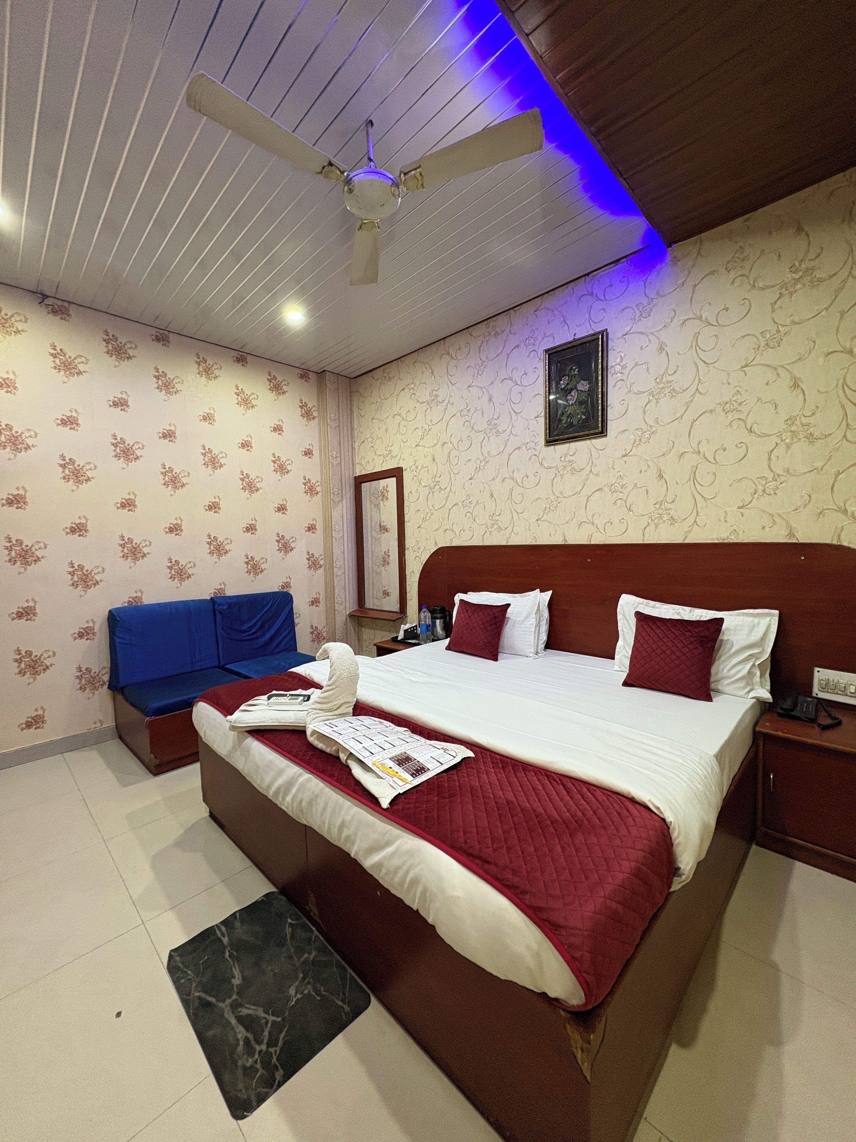 Camera d'albergo con un letto grande, un divano blu, una ventola a soffitto e un tavolino con un telefono e una bottiglia.