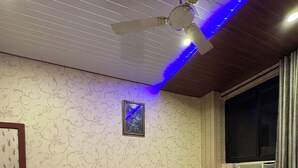 Ein Hotelzimmer mit einem großen Bett, einem Deckenventilator, einem Nachttisch, einem Fenster mit Vorhängen und einem an der Wand befestigten Bild.