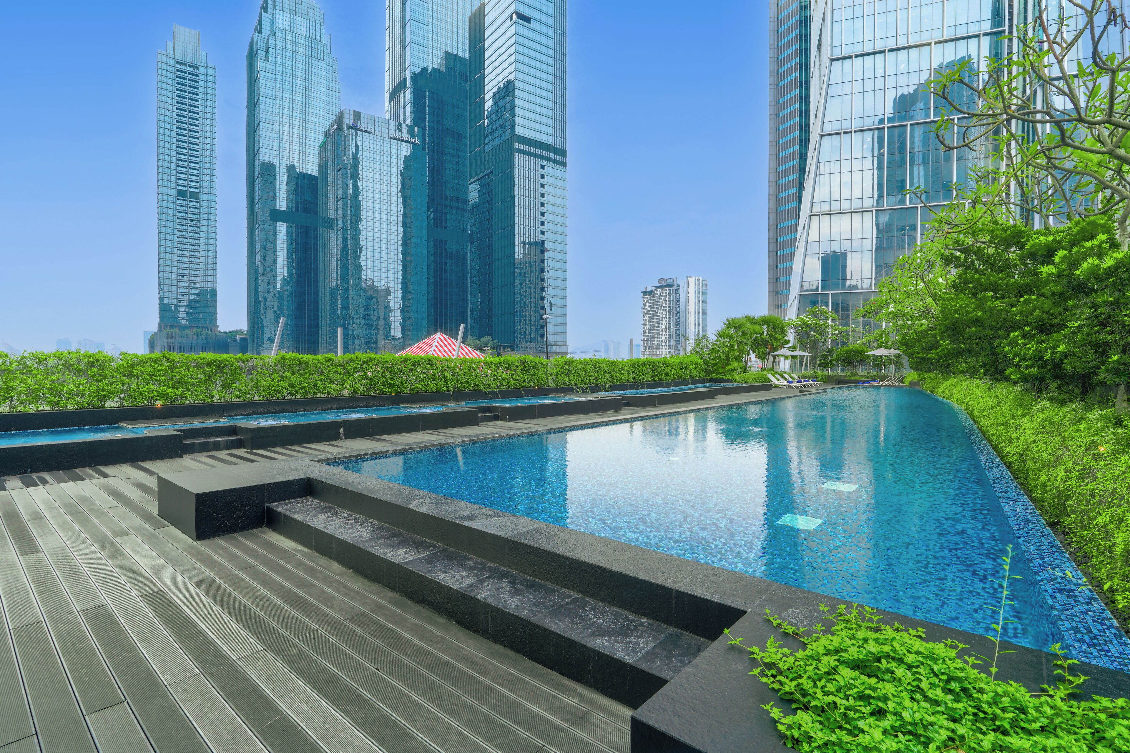 Ein moderner Pool mit Holzterrasse, umgeben von üppiger Vegetation und hohen Gebäuden im Hintergrund.