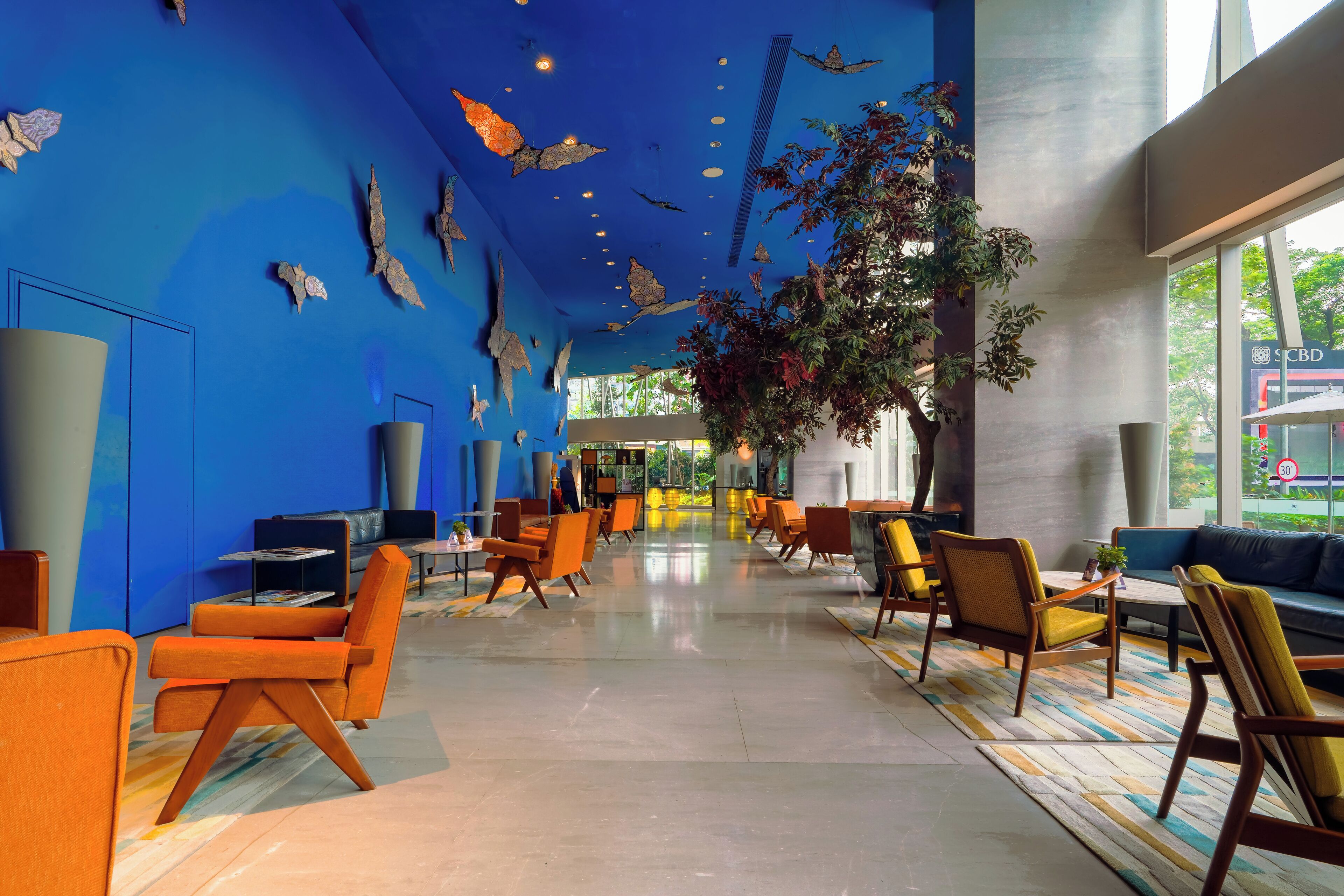 A spacious lobby with modern orange and yellow seating, a large indoor plant, and a blue ceiling with fish wall art.