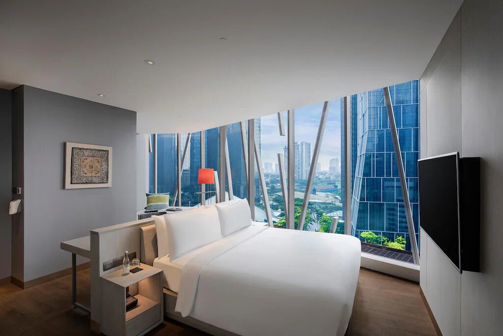 Habitación de hotel moderna con una cama grande, una mesita de noche, un televisor y vistas a la ciudad a través de ventanales del suelo al techo.