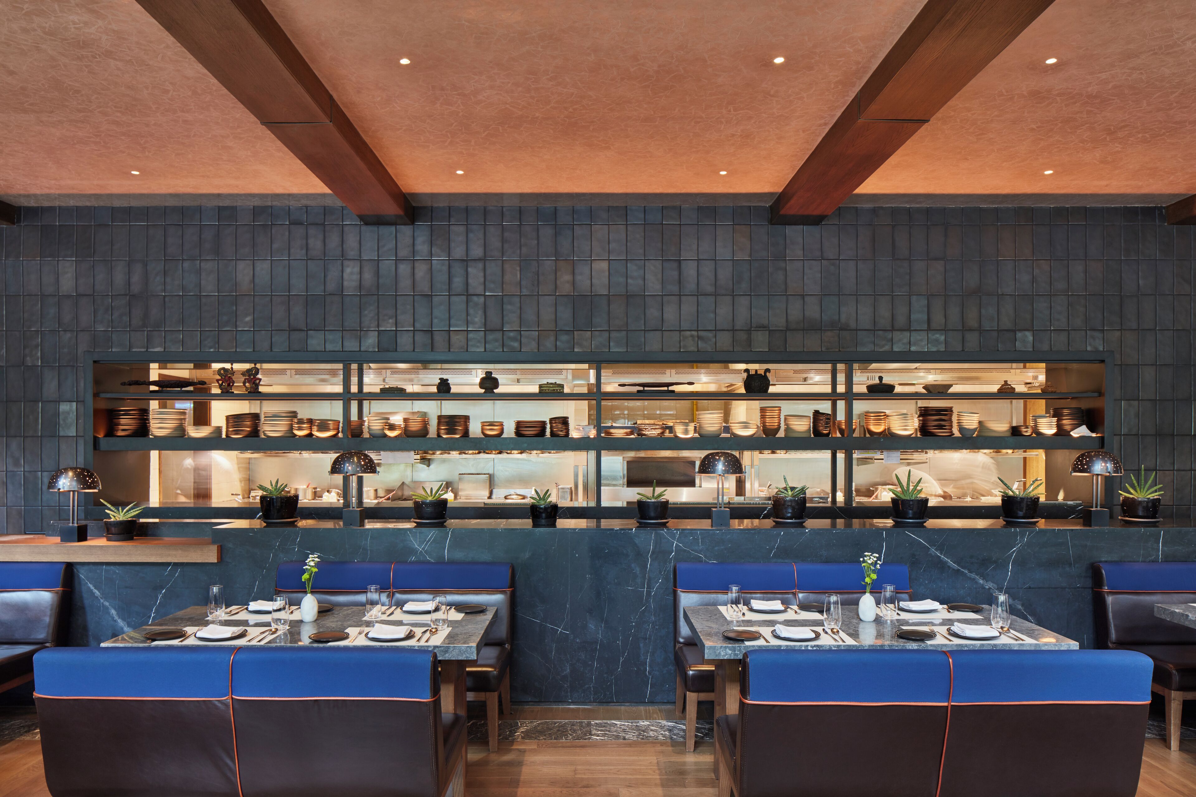 Un restaurante con mesas de mármol y asientos tapizados en azul, una zona de cocina con una exhibición de platos, y un diseño de techo moderno.