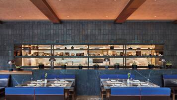 Un restaurante con mesas de mármol y asientos tapizados en azul, una zona de cocina con una exhibición de platos, y un diseño de techo moderno.