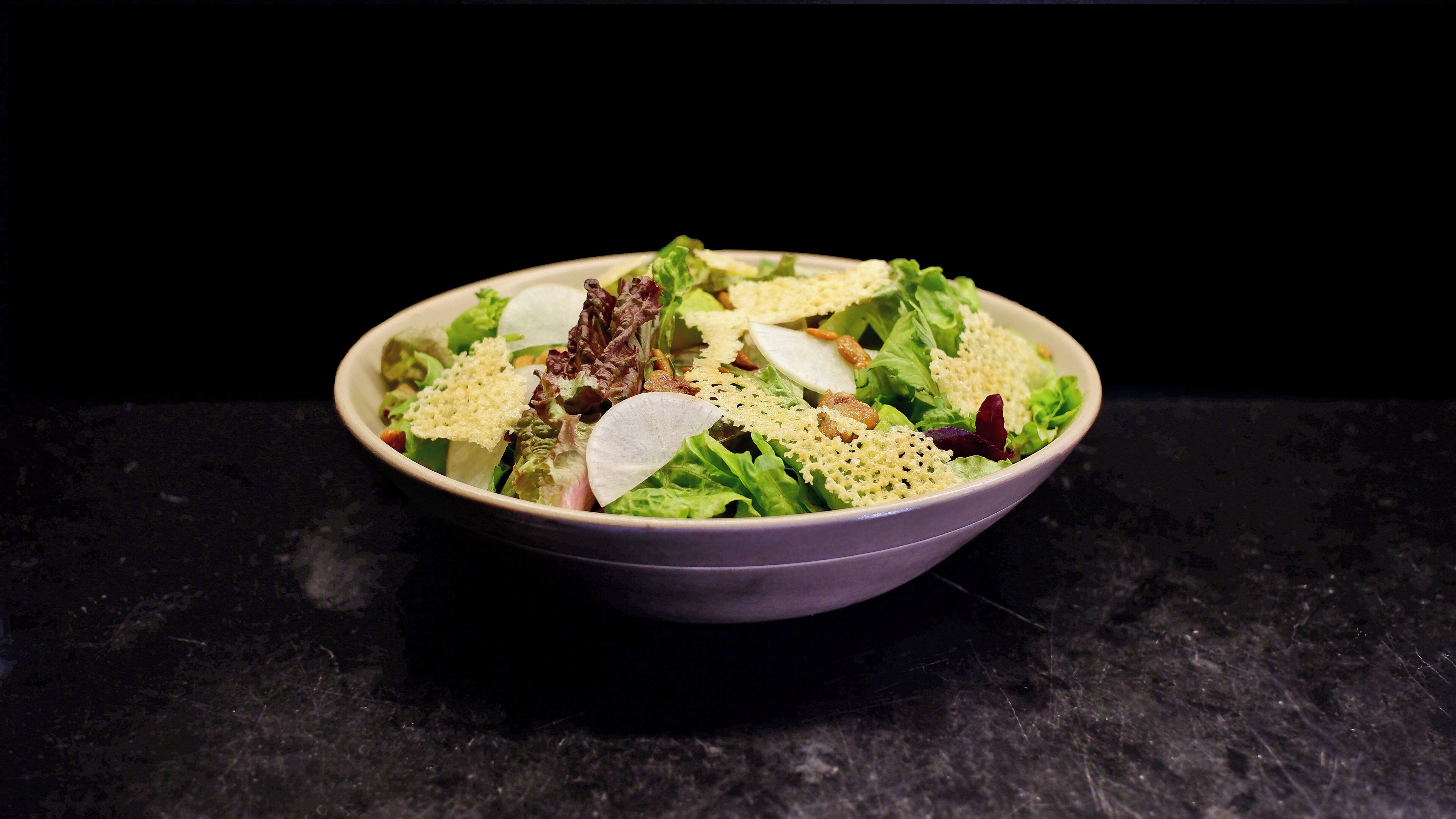 Un saladier rempli de salade, avec différentes sortes de légumes verts et des croûtons, posé sur une surface sombre.