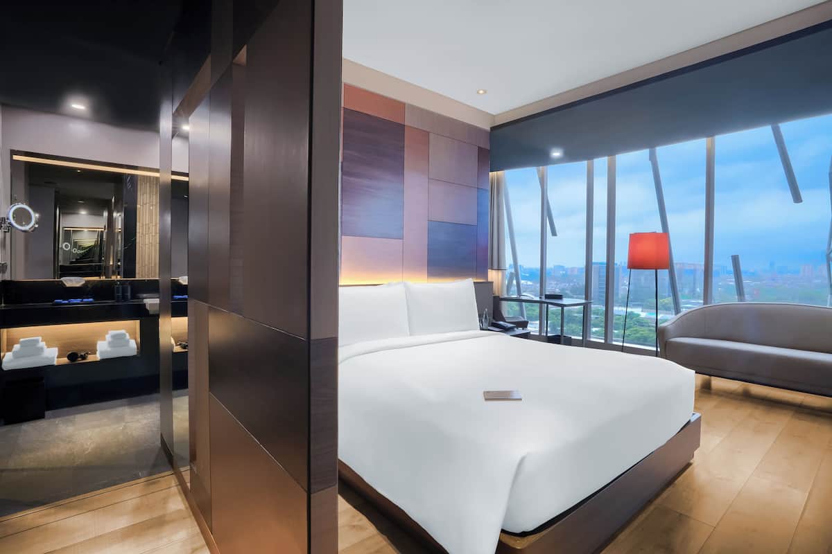 Una habitación de hotel moderna con una cama grande, un escritorio con computadora, un tocador con espejo y un ventanal con vista a la ciudad.