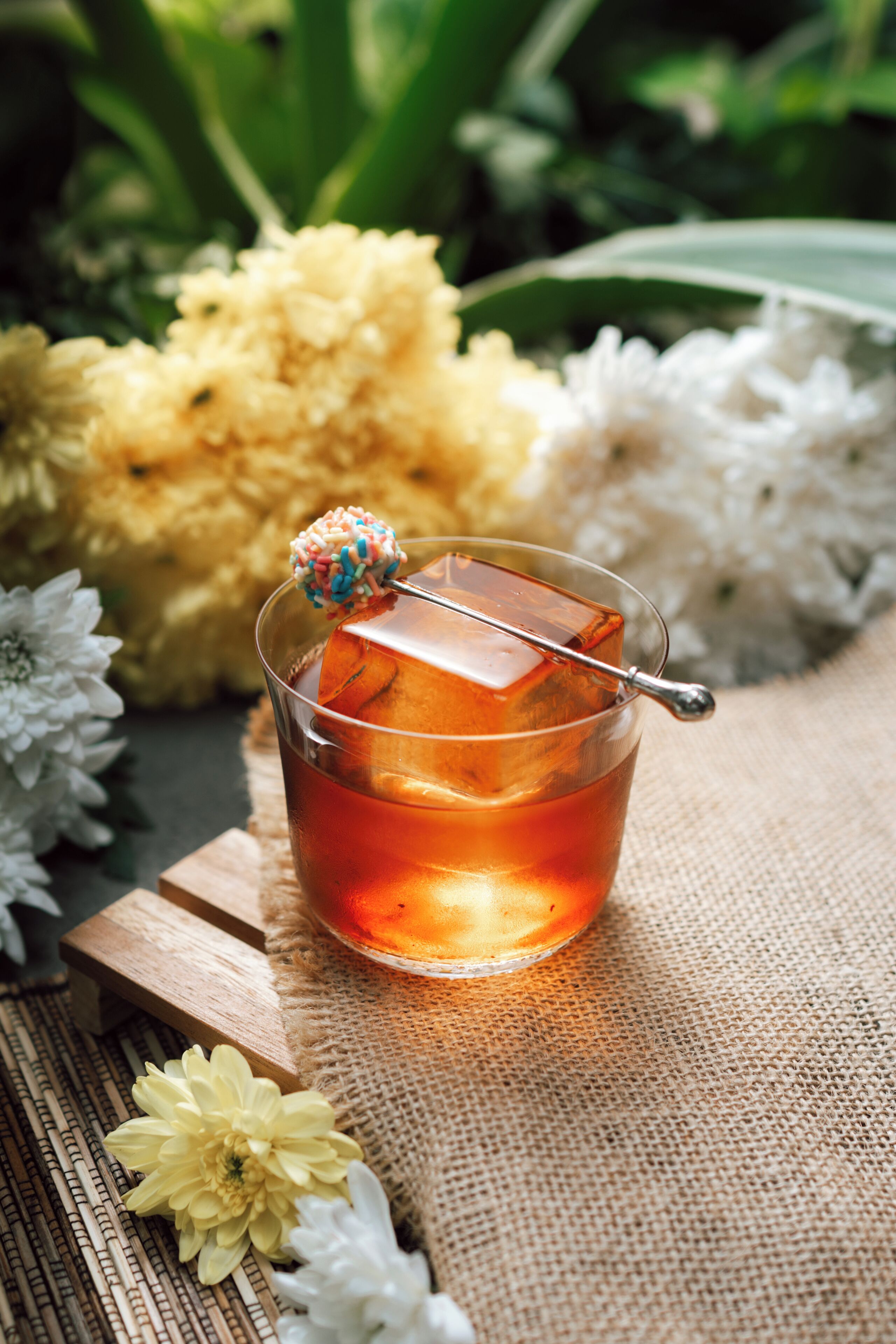 A clear glass with a cube-shaped ice cube and a colorful candy on a stick, placed on a wooden surface with flowers and a burlap cloth.
