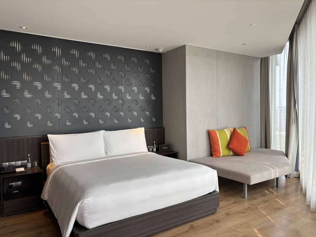 A modern hotel room with a large bed, a bedside table, a nightstand, a sofa, and a patterned wall.