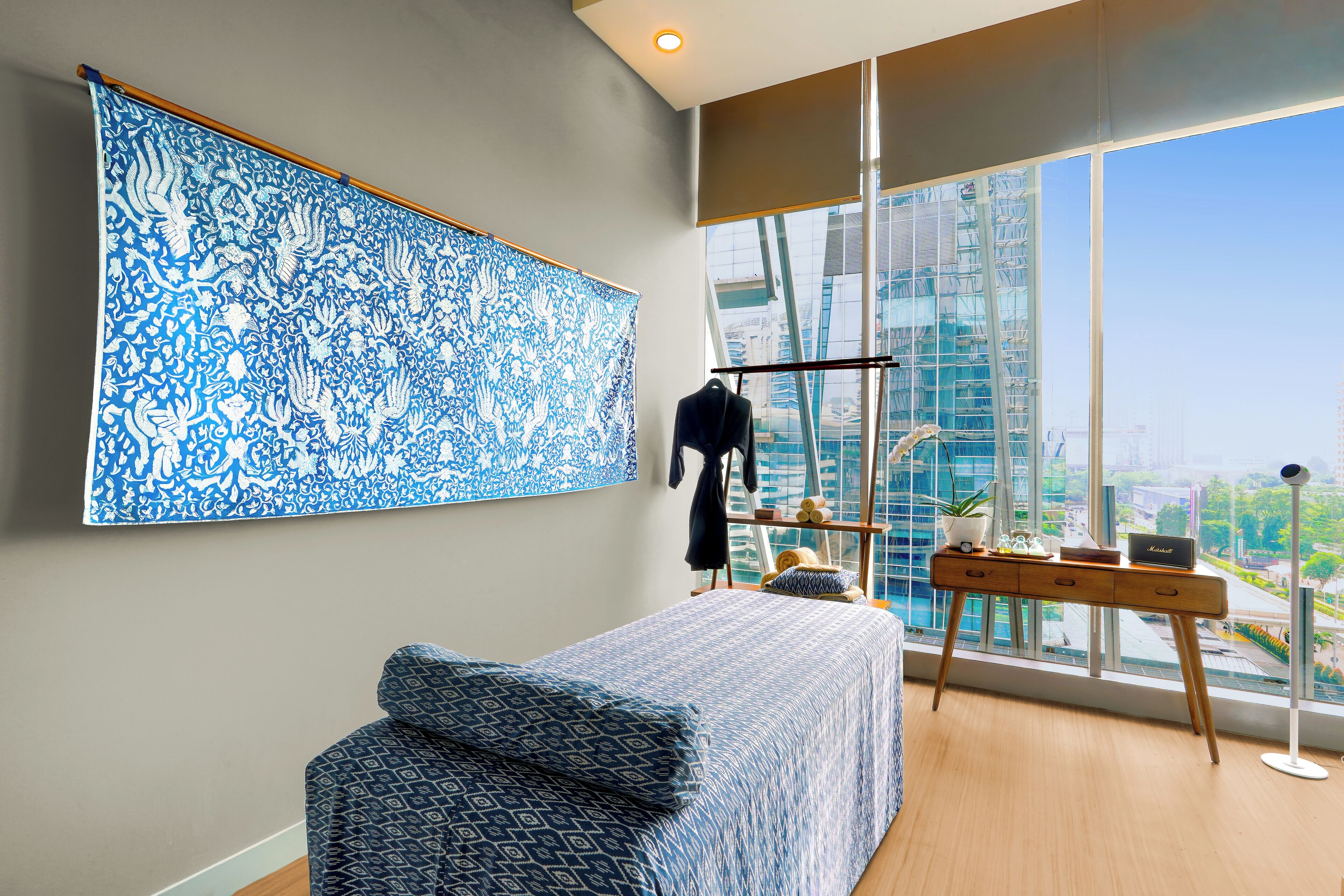 Une chambre moderne avec une grande fenêtre, un lit recouvert d’une literie à motifs, un bureau avec un ordinateur portable et une tapisserie décorative.