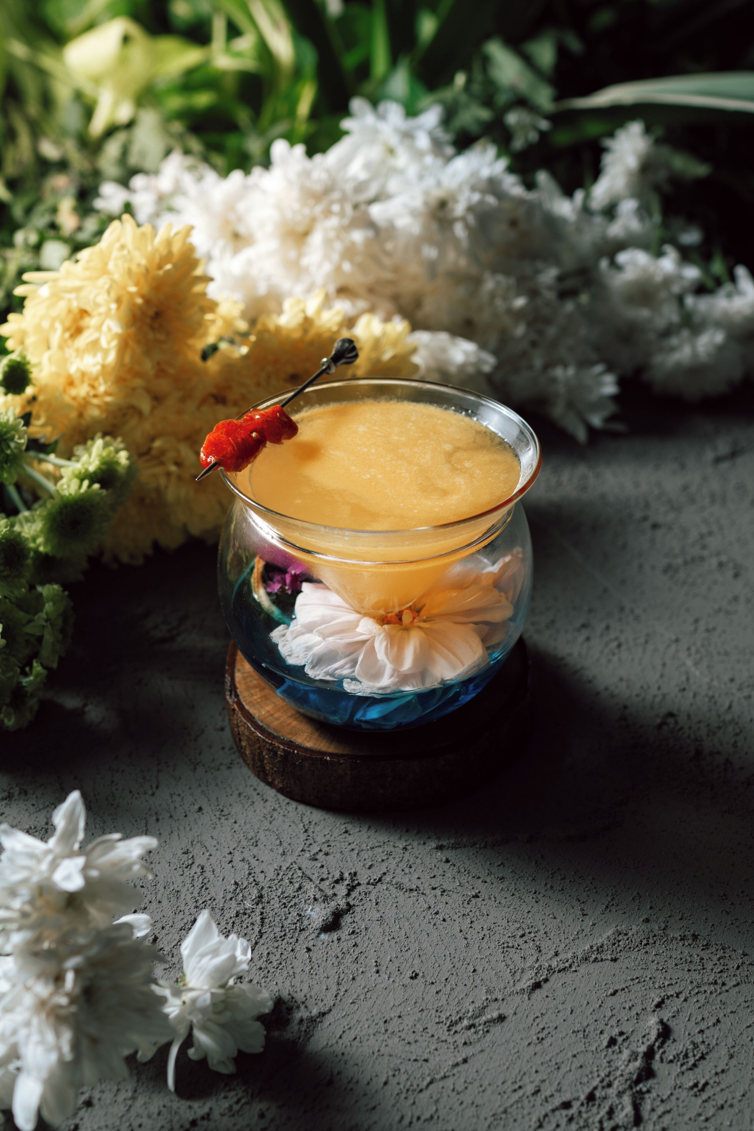 A cocktail in a glass with a red straw, surrounded by flowers on a textured surface.