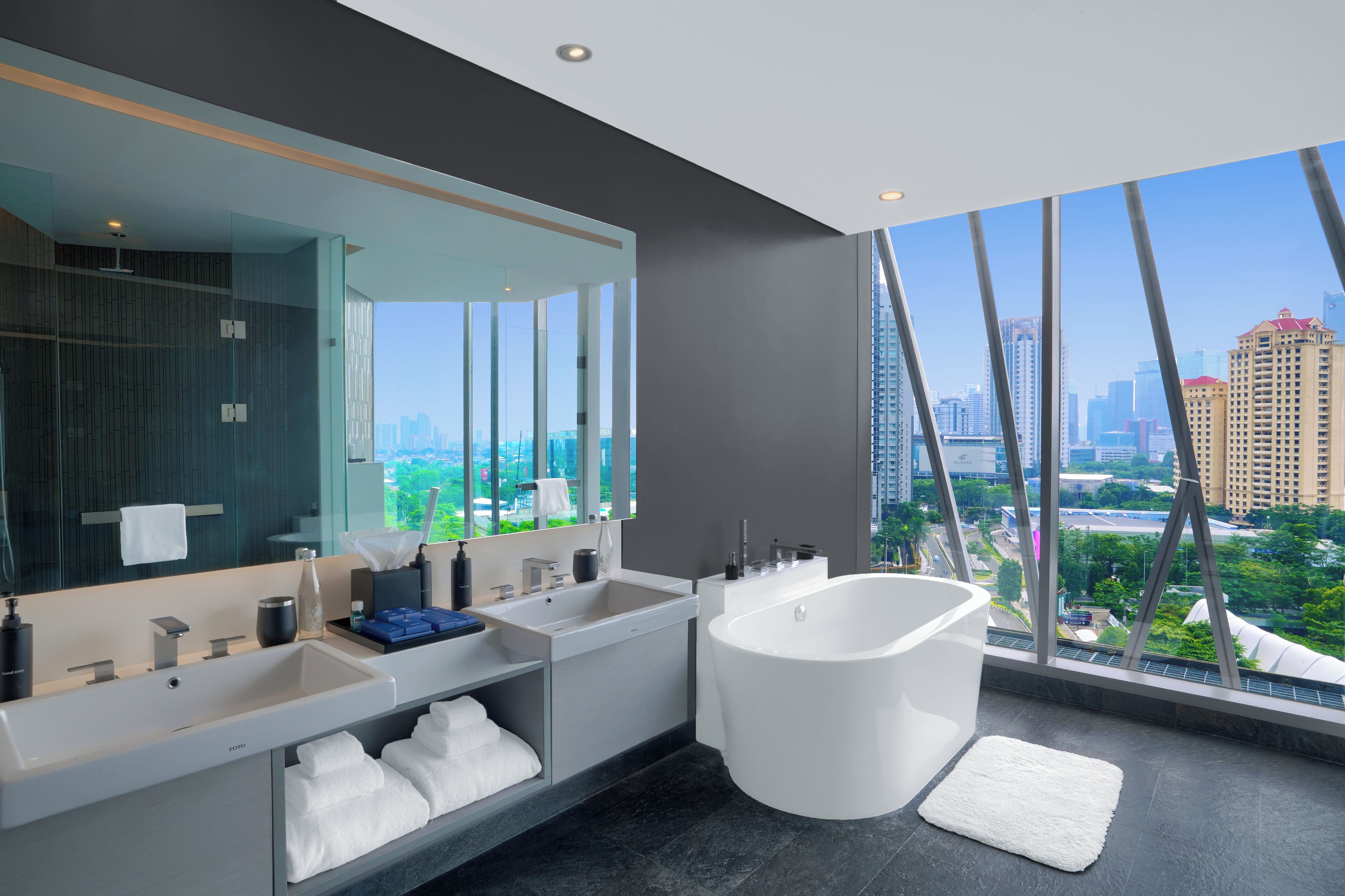 A modern bathroom with a large bathtub, two sinks, and a large mirror. It features a city view through large windows.