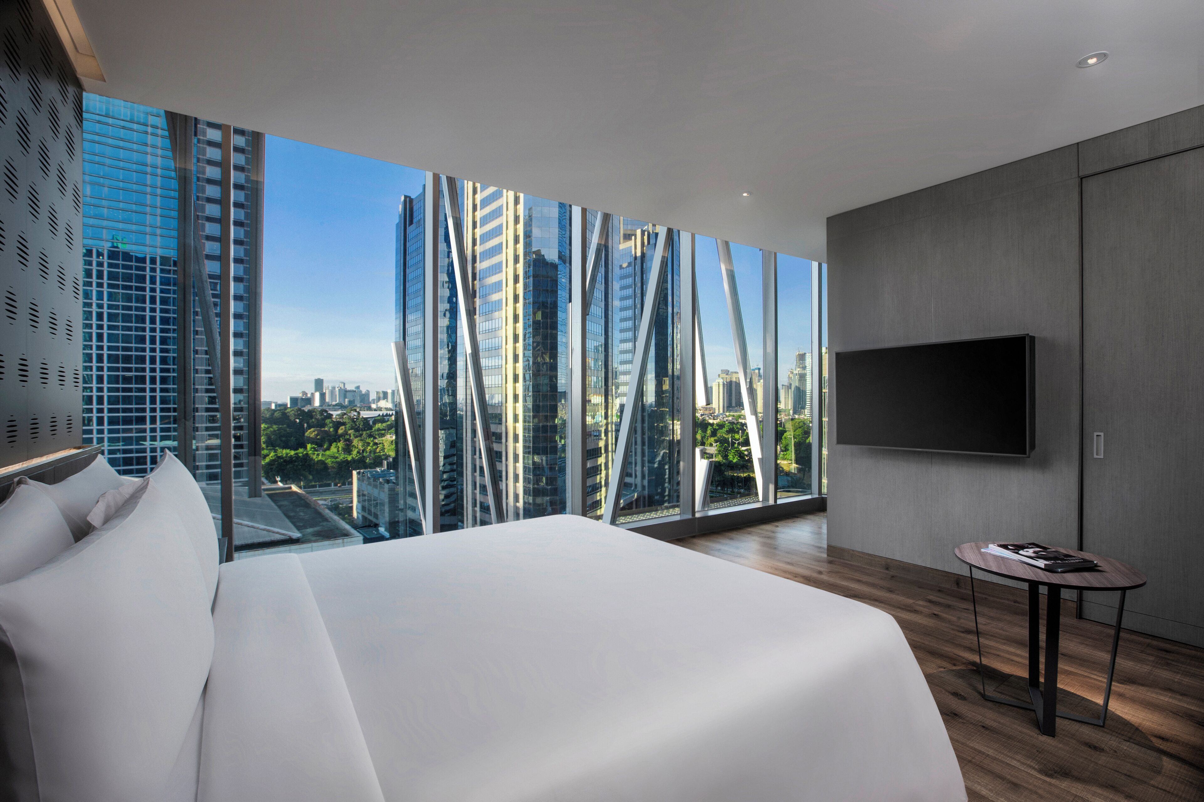 A modern hotel room with a large bed, a TV mounted on the wall, and a view of tall buildings through floor-to-ceiling windows.