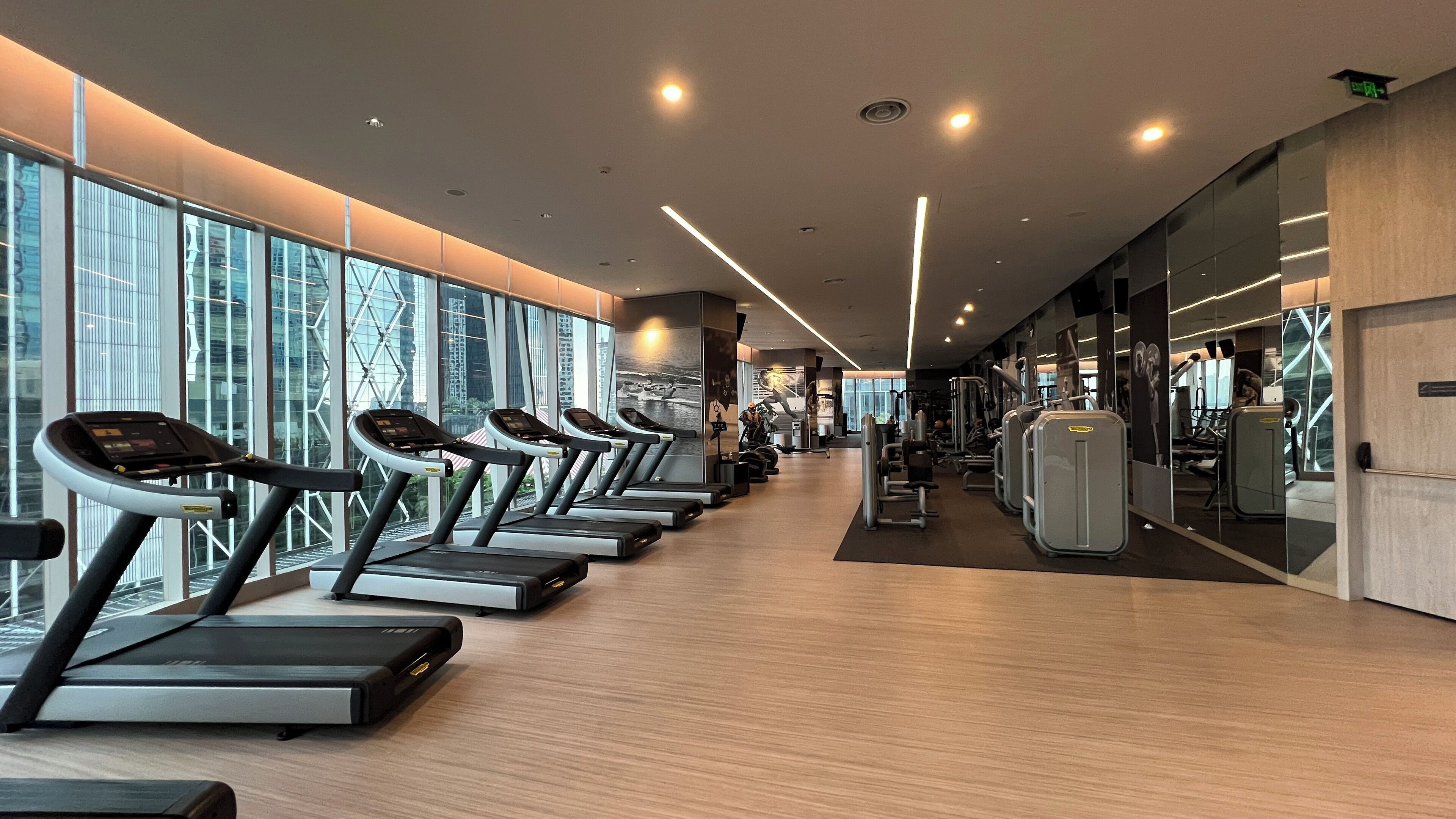 Un gimnasio moderno con cintas de correr alineadas a lo largo de una pared con ventanas amplias, y diversas máquinas de ejercicio al fondo.