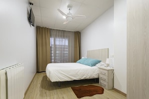 Un dormitorio con cama, mesita de noche, lámpara, ventilador de techo, ventana con cortinas y radiador.