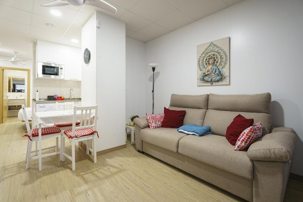 A living room with a sofa, a dining table with chairs, a kitchen area, and a wall-mounted artwork.