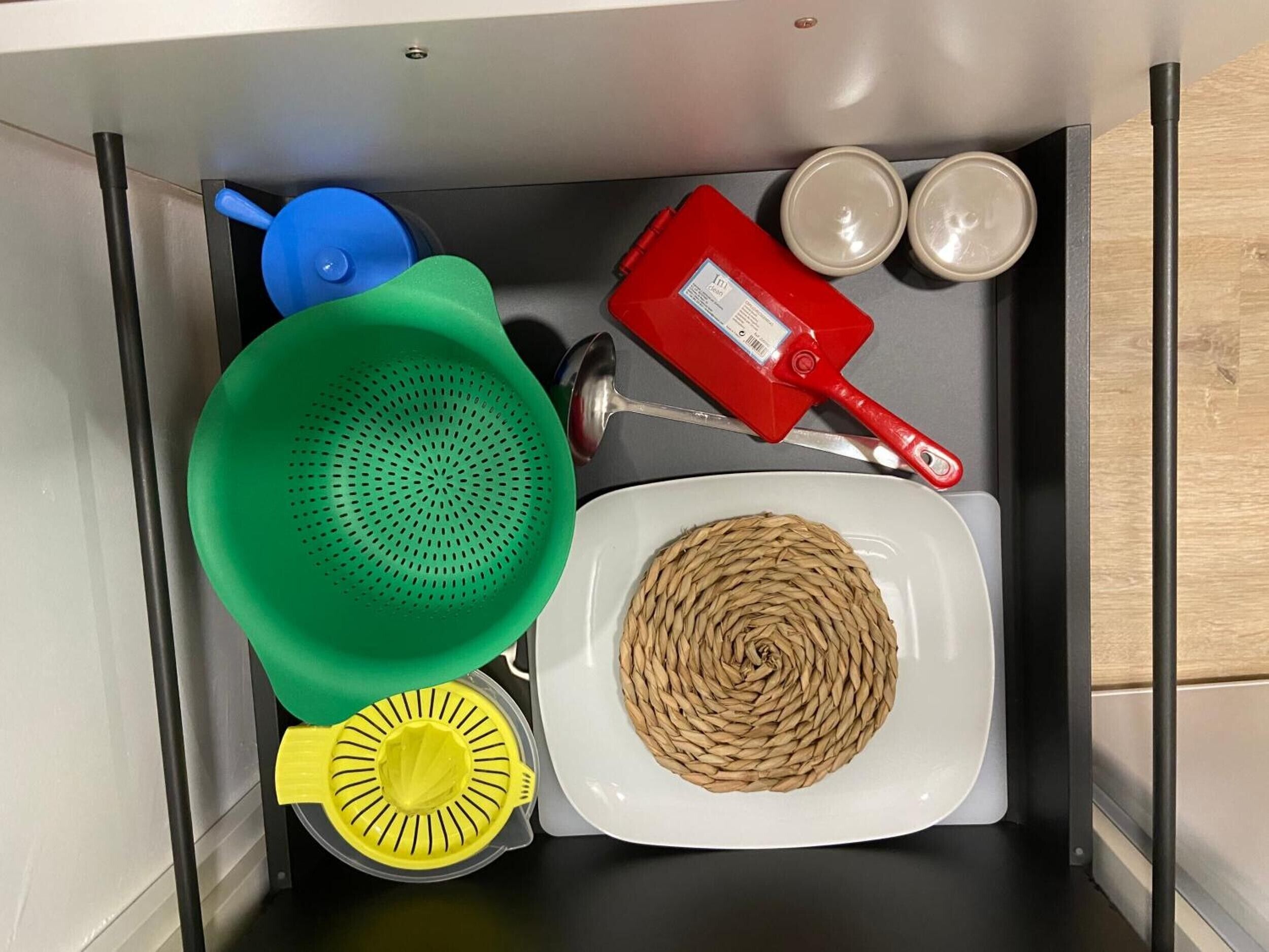 Un cajón de cocina con un colador, un exprimidor, un batidor, una botella roja y dos recipientes pequeños.