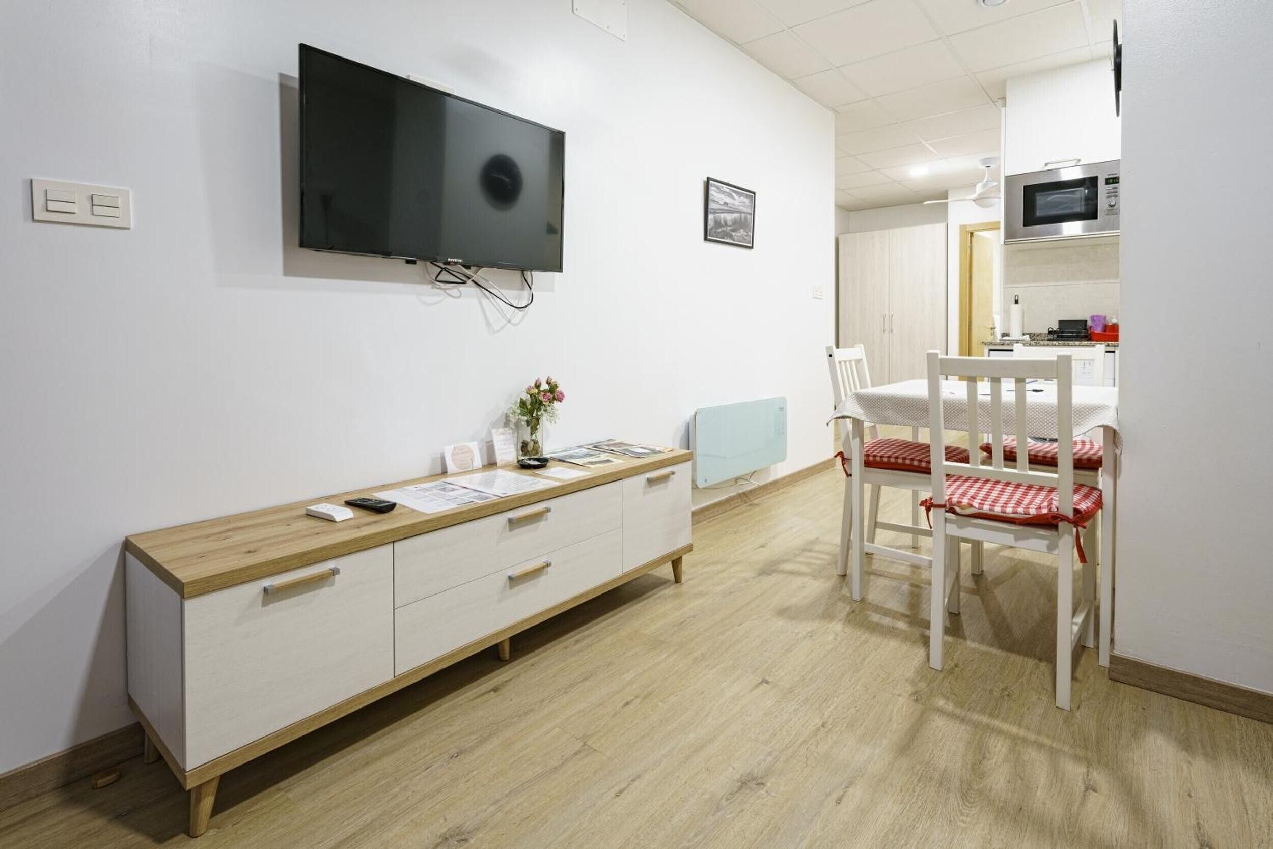 A room with a TV mounted on the wall, a wooden cabinet, a dining table with chairs, and a kitchen area in the background.